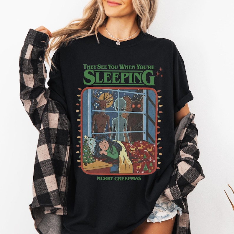 Op de afbeelding: Een zwart t-shirt met een grafische print met de tekst "THEY SEE YOU WHEN YOU'RE SLEEPING" en "MERRY CREEPMAS". De illustratie toont drie wezens die door een raam naar een persoon in bed kijken, omringd door kerstverlichting en decoraties.