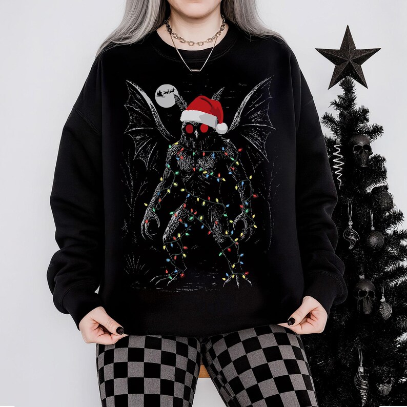 K&ouml;nnte beinhalten: Schwarzes Sweatshirt mit einem Mothman-Motiv, das einen Weihnachtsmannhut und Lichterketten tr&auml;gt. Das Design umfasst einen Vollmond und einen Weihnachtsbaum mit Totenkopf-Dekorationen. Die Person tr&auml;gt eine karierte Hose.
