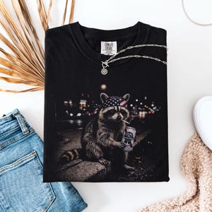 K&ouml;nnte beinhalten: Schwarzes Comfort Colors T-Shirt mit einem Waschb&auml;ren, der ein amerikanisches Flaggen-Bandana tr&auml;gt, raucht und eine Bierdose h&auml;lt. Eine silberne Halskette liegt auf dem Shirt. Das Shirt wird mit blauen Jeans und einem beigen Pullover kombiniert.