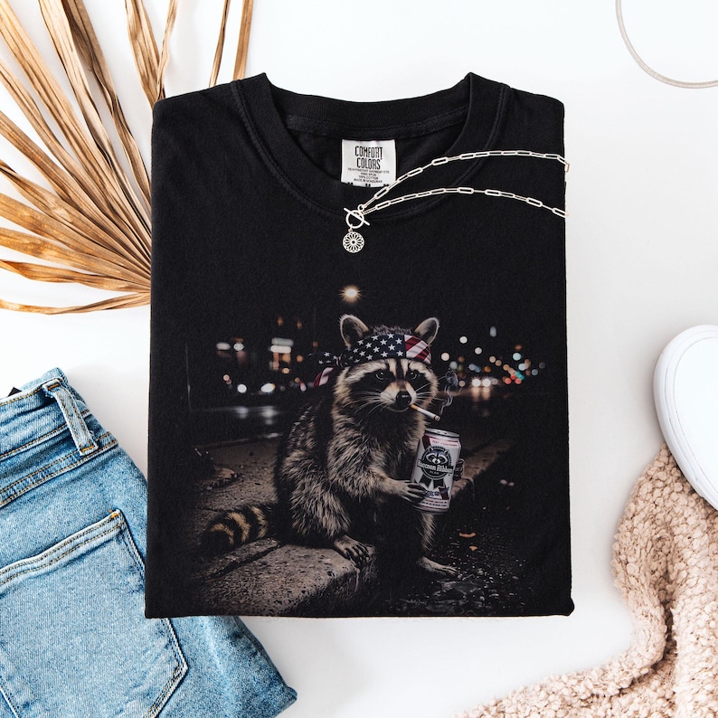 K&ouml;nnte beinhalten: Schwarzes Comfort Colors T-Shirt mit einem Waschb&auml;ren, der ein amerikanisches Flaggen-Bandana tr&auml;gt, raucht und eine Bierdose h&auml;lt. Eine silberne Halskette liegt auf dem Shirt. Das Shirt wird mit blauen Jeans und einem beigen Pullover kombiniert.