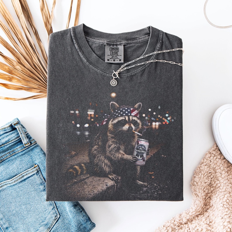 K&ouml;nnte beinhalten: Dunkelgraues Comfort Colors T-Shirt mit einem Waschb&auml;ren, der ein Bandana mit amerikanischer Flagge tr&auml;gt, eine Zigarette raucht und eine Bierdose h&auml;lt. Das T-Shirt hat einen Rundhalsausschnitt und kurze &Auml;rmel. Eine silberne Halskette liegt &uuml;ber dem T-Shirt.