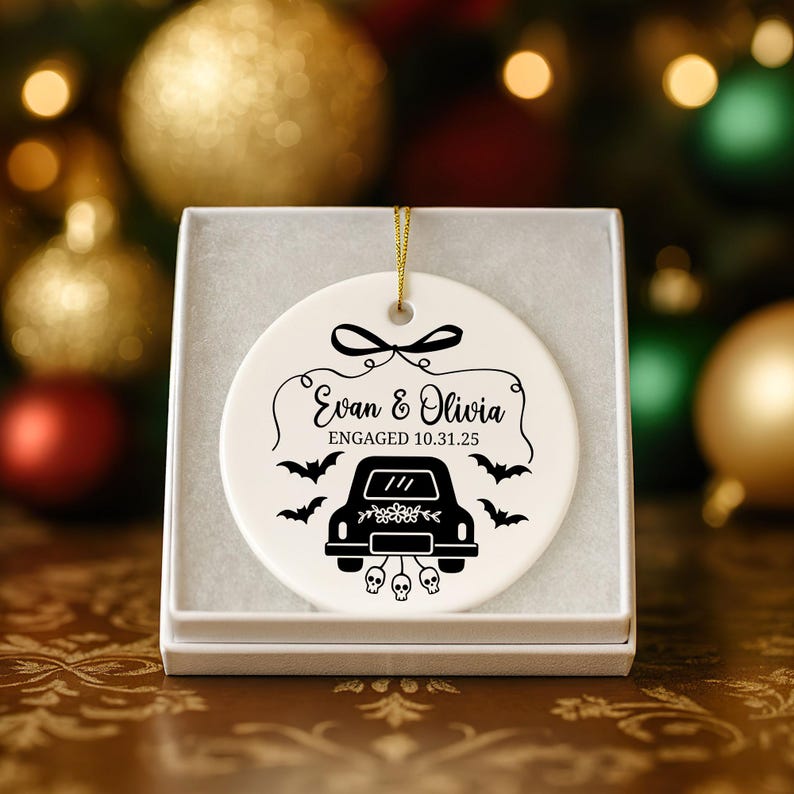 May include: A white ceramic ornament with a black car graphic, bats, and the text "Evan & Olivia Engaged 10.31.25." The ornament is in a white gift box, perfect for engagement celebrations.