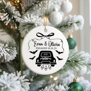 May include: White ceramic Christmas ornament with black text and graphics. The ornament features the names "Evan & Olivia" and the date "10.31.25" with a car, bats, and skulls design. A gold string is attached for hanging.