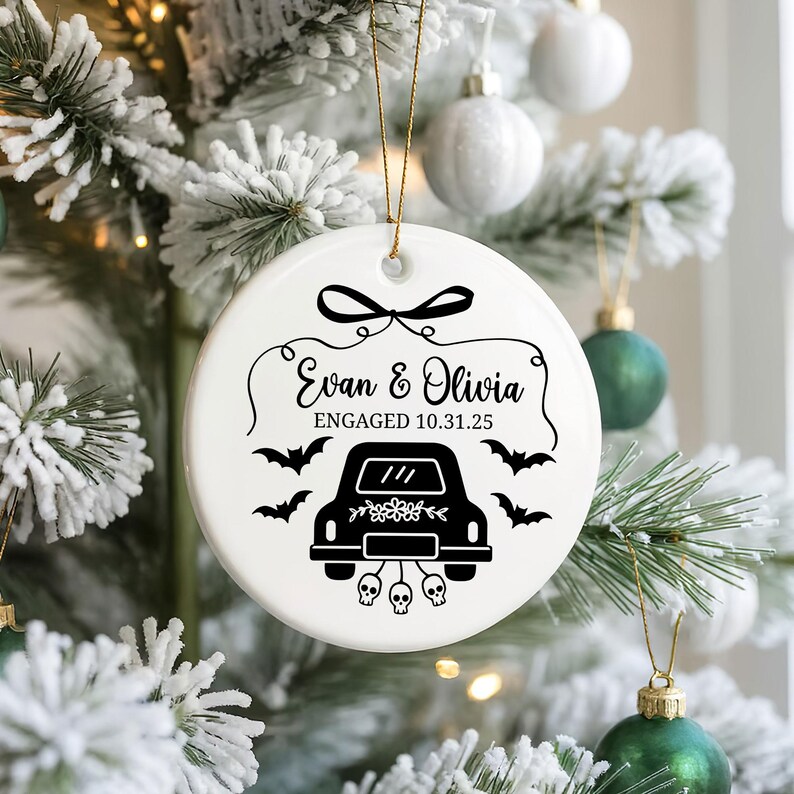 May include: White ceramic Christmas ornament with black text and graphics. The ornament features the names "Evan & Olivia" and the date "10.31.25" with a car, bats, and skulls design. A gold string is attached for hanging.