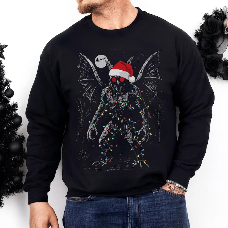 K&ouml;nnte beinhalten: Schwarzes Sweatshirt mit einem Aufdruck eines Mothman-Kreatur, die einen Weihnachtsmannhut und Lichterketten tr&auml;gt. Das Design beinhaltet einen Vollmond und den Schlitten des Weihnachtsmanns. Die Kreatur hat rote Augen und fledermausartige Fl&uuml;gel.