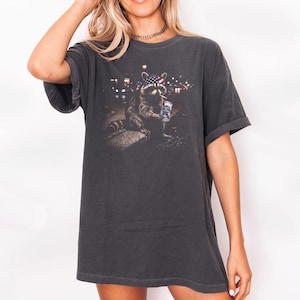 K&ouml;nnte beinhalten: Dunkelgraues T-Shirt mit einem Waschb&auml;r-Motiv, das ein Bandana mit amerikanischem Flaggenmuster tr&auml;gt und eine Dose h&auml;lt. Das Bild hat eine n&auml;chtliche Stadtszene. Das T-Shirt hat kurze &Auml;rmel und eine lockere Passform.