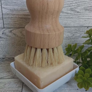 Op de afbeelding: Een afwasborstel met houten handvat en natuurlijke borstelharen rust op een stuk zeep in een witte, vierkante zeepbakje. De zeep is lichtbeige. De borstelsteel is lichtbruin.