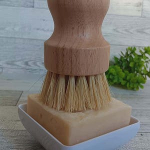 Può includere: Una spazzola per piatti con manico in legno e setole naturali poggia su una saponetta all'interno di un portasapone bianco quadrato. La spazzola ha una parte superiore arrotondata e le setole sono di colore marrone chiaro. Il sapone è beige chiaro.