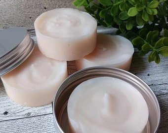 May include: Four round, white, creamy soap bars in various containers. Three bars are stacked on top of each other, and the fourth bar is in a silver tin.