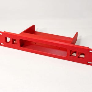 Può includere: Un componente per montaggio a rack rosso brillante con aperture rettangolari e circolari. Il componente ha una base piatta con supporti verticali e fori di montaggio. Il design è pulito e moderno.