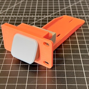 May include: An orange and gray 3D-printed device with a rectangular base and a light gray square component. The device has a flat, angled top and is set against a black grid background.