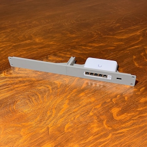 May include: A gray network rack mount with a white device and multiple ports. The white device has a curved logo. The rack mount is rectangular with a series of ports and a small rectangular opening. The background is a wooden surface.