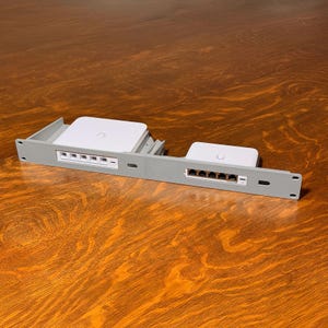 May include: A gray rack mount with two white network devices. Each device has a curved logo. The rack has multiple ports and is designed to hold the devices securely. The devices are likely used for networking or data transfer.