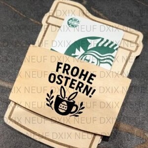 Puede incluir: Un tarjetero de madera con forma de taza de café, con una tarjeta de regalo de Starbucks en su interior. El soporte es de color marrón claro y presenta el texto "FROHE OSTERN!" y un diseño de conejito.