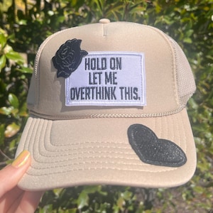 May include: A beige trucker hat with a black rose patch, a black heart patch, and a white patch that says "Hold on let me overthink this."