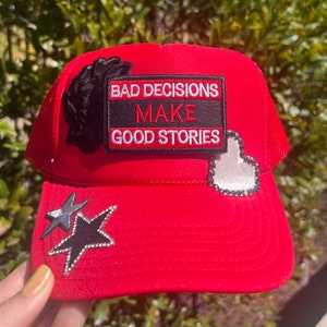 May include: Red trucker hat with a black and white patch that says "Bad Decisions Make Good Stories". The hat also has a black star with rhinestones, a white heart with rhinestones, and a black flower with rhinestones.