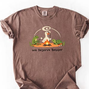 May include: Brown t-shirt featuring a cartoon frog, duck, and turtle gathered around a campfire, with the words "We Deserve Better". The tee has a vintage, faded appearance.