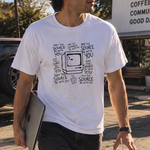 May include: A white short-sleeved t-shirt featuring a black graphic of a vintage computer monitor surrounded by the repeated text "May the SOURCE be with you". The shirt is paired with dark grey denim jeans and white sneakers.