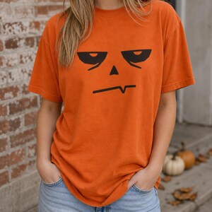 May include: An orange short-sleeved t-shirt featuring a black graphic print of a stylised, grumpy pumpkin face with heavy-lidded eyes, a small triangular nose, and a jagged, uneven mouth line centred on the chest area of the garment.