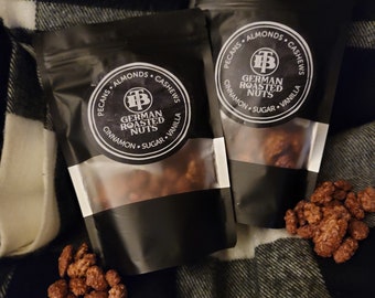 May include: Two black bags of German roasted nuts with a white label that says "German Roasted Nuts" with a logo of a capital "T" and a capital "B" inside a circle. The label also says "Pecans, Almonds, Cashews, Cinnamon, Sugar, Vanilla". The bags are on a black and white plaid surface.