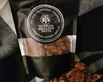 Könnte beinhalten: Eine schwarze Tüte mit einem transparenten Fenster, in dem verschiedene geröstete Nüsse zu sehen sind. Die Tüte hat ein weißes Etikett mit dem Text "German Roasted Nuts" und einer Zutatenliste: Pekannüsse, Mandeln, Cashews, Zimt, Zucker und Vanille.