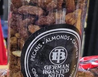May include: A clear glass jar filled with a variety of German roasted nuts. The jar has a black lid and a black and white label that reads "German Roasted Nuts" with the ingredients listed around the label: pecans, almonds, cashews, cinnamon, sugar, vanilla.