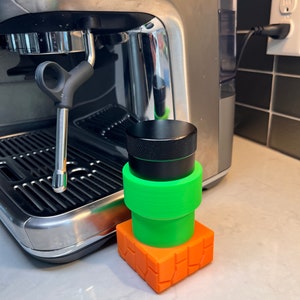 May include: A black and green coffee grinder sits on top of an orange and black platform. The grinder is sitting next to a silver espresso machine.