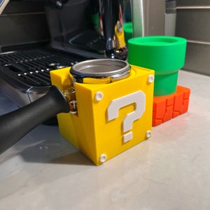 May include: A yellow 3D printed question mark block with a white question mark, designed to hold a coffee tamper. The block is sitting on a countertop next to a green and orange pipe-shaped object.