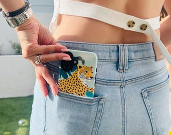 May include: A phone case with a blue background and a yellow and black leopard print. The leopard is surrounded by green leaves and pink, yellow, and blue flowers.