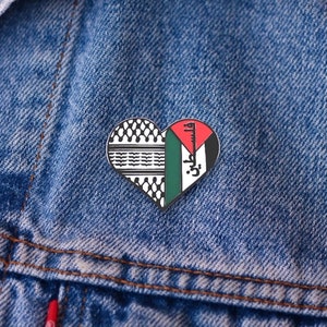 May include: Heart-shaped pin with a design split into two halves. One side features a black and white pattern, the other side displays the Palestinian flag with Arabic text. The pin is set against a blue denim background.
