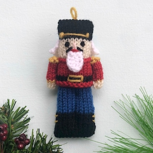 Peut inclure: Une décoration de Noël en forme de casse-noisette tricotée avec un uniforme rouge et bleu, un chapeau noir, une barbe blanche et des accents dorés.