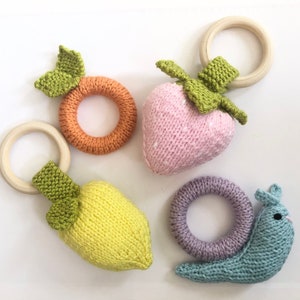 May include: Four knitted baby teethers in the shape of a lemon, strawberry, snail, and orange. The teethers are attached to wooden rings.