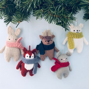 May include: Five knitted animal decorations hanging from a pine tree branch. The decorations are a white bunny with pink ears and a pink scarf, a brown reindeer with a blue scarf, a red fox with a grey scarf, a white polar bear with a yellow scarf, and a grey bear with a red scarf.