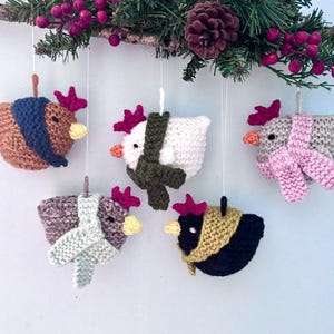 May include: Five crocheted chicken ornaments hanging from a branch with pine needles and red berries. Each chicken features a unique body color, a red comb, a yellow beak, black eyes, and a knitted scarf in a contrasting color.