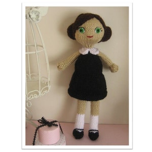May include: A knitted doll with brown hair, green eyes, and a black dress. The doll is wearing white socks and black shoes. The doll is standing on a pink surface.