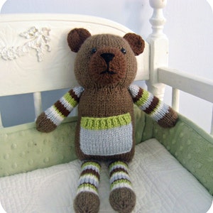 May include: A brown knitted teddy bear with green and white striped arms and legs. The bear is wearing a light blue and green knitted vest. The bear is sitting on a white crib with a green and white patterned bumper.