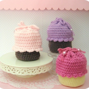 May include: Three crocheted cupcake toys on white cake stands. The cupcakes are pink, purple, and yellow with brown frosting. The cupcakes are decorated with white beads.