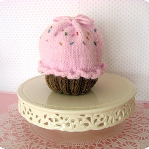 May include: A pink knitted cupcake hat with brown frosting and colorful sprinkles. The hat is sitting on a white cake stand with a lace doily underneath.