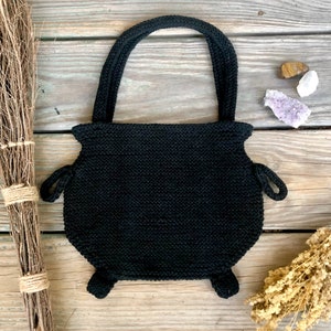 May include: Black knitted cauldron-shaped tote bag with a black handle. The bag is sitting on a wooden surface.