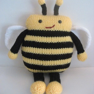 Puede incluir: Un juguete de abeja de punto con rayas negras y amarillas, alas blancas y antenas negras. La abeja tiene una cara sonriente con ojos negros y una boca roja.