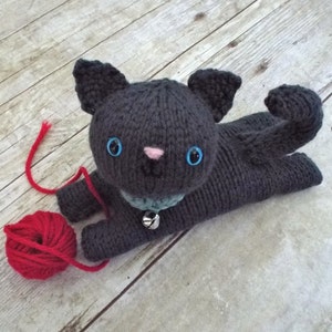 May include: A gray knitted cat toy with blue eyes and a pink nose. The cat is holding a red ball of yarn.