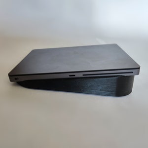 May include: A dark grey laptop on a black laptop stand. The stand is angled to raise the laptop, and is made of a textured material. The laptop is closed, with a sleek, modern design. The stand is designed to improve ergonomics.