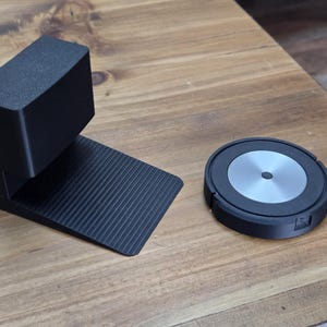 May include: Black robotic vacuum cleaner and a black charging dock on a wooden surface. The dock has a rectangular base with a ridged surface and an upright section. The vacuum is round with a silver center.