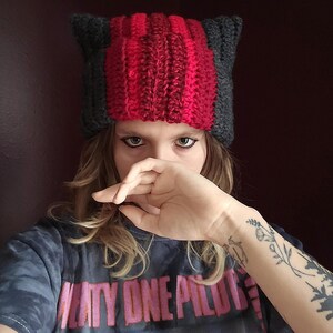May include: A handmade cat ear beanie in a ribbed knit pattern. The hat features a central section of vibrant red yarn flanked by gray sections, with matching gray ears. The beanie is a unique, cozy accessory.