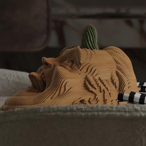 Puede incluir: Una escultura de madera de una cara con un pequeño cactus que crece desde la parte superior. Una botella de vino oscuro y dos copas de vino están sobre la mesa. La escultura está sobre una mesa con un paño a rayas blancas y negras.