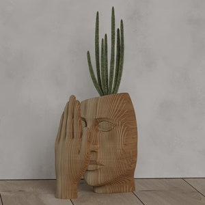 Op de afbeelding: Een houten bloempot in de vorm van een gezicht met een hand die de mond bedekt. De pot bevat een groene plant. Het hout heeft een natuurlijk nerfpatroon. Een uniek decoratief item.