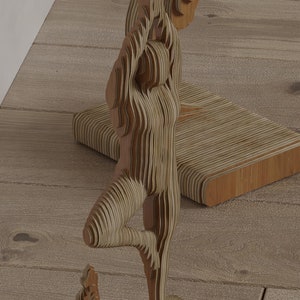 Puede incluir: Escultura de madera de una persona en postura de yoga, con los brazos levantados por encima de la cabeza y una pierna levantada. La escultura está hecha de madera en capas, creando un efecto tridimensional. El color es marrón claro.