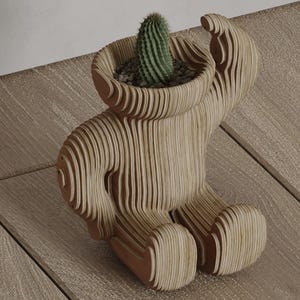 May include: A wooden bear-shaped planter with a cactus inside. The planter is made of layered wood, creating a textured effect. The cactus is green and spiky, and the planter is sitting on a wooden surface.