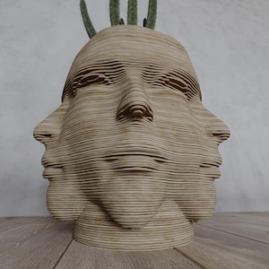 Puede incluir: Escultura de madera en capas de color marrón claro que representa un rostro humano, con múltiples caras visibles. La escultura tiene un aspecto texturizado y en capas. Pequeños cactus verdes plantados en la parte superior. La escultura está sobre una superficie de madera.