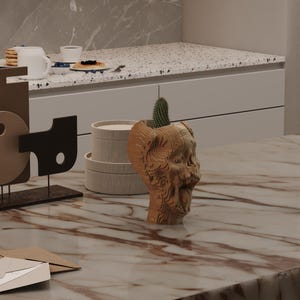Puede incluir: Una maceta de madera en forma de cabeza, de color marrón claro, con un pequeño cactus en su interior. La maceta tiene un diseño tallado detallado. Se encuentra sobre una encimera de mármol, con otros objetos decorativos en el fondo.